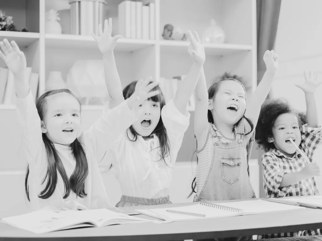Four children sitting at a desk with open books, raising their hands and smiling in a classroom setting.