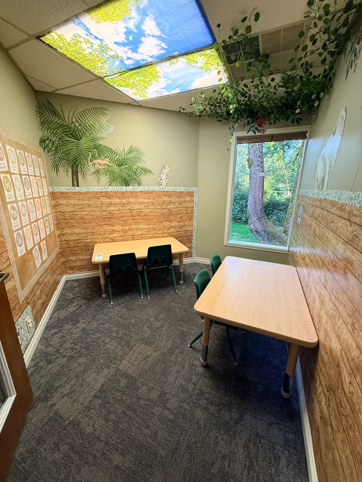 Classroom with tropical jungle mural and blue sky ceiling, tables, and chairs.