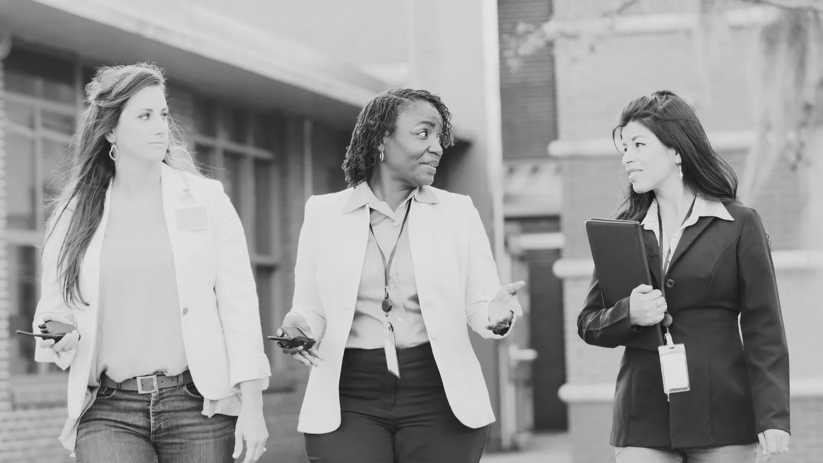 Three women walking and talking outdoors, two holding phones and one holding a notebook, dressed in business casual attire.