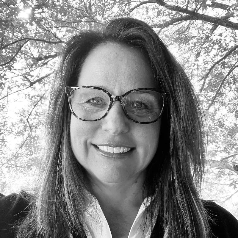 A woman with long dark hair wearing glasses with a patterned frame, smiling outdoors with trees and sunlight in the background.