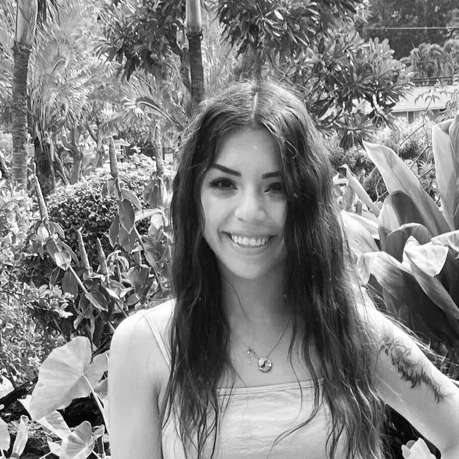 A woman with long wavy hair smiling outdoors among lush tropical plants. She is wearing a sleeveless top and a necklace with two pendants.