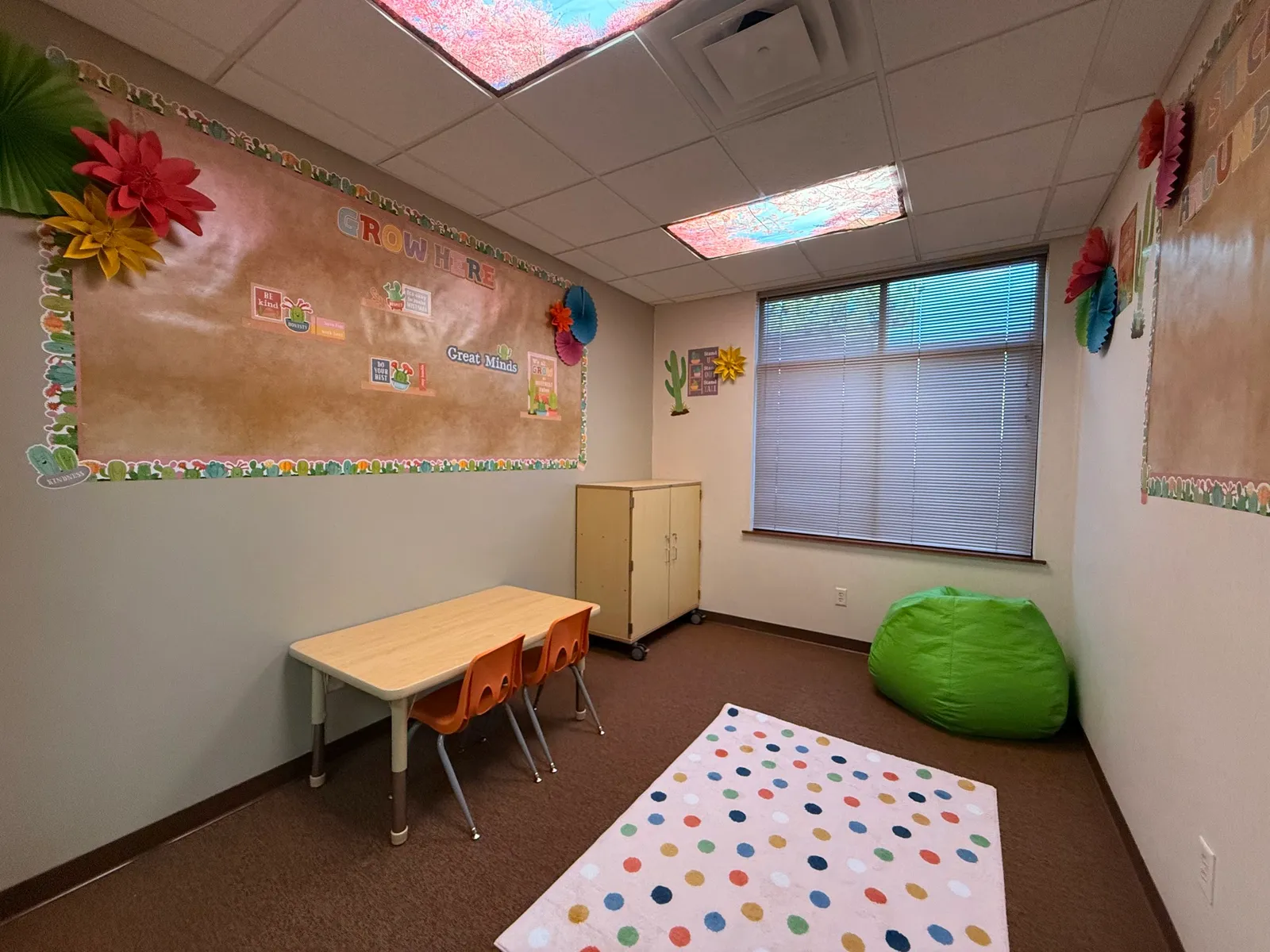 A small classroom corner with a beige wall decorated with a bulletin board titled GROW HERE and colorful paper flowers, a window with blinds, a small wooden table with four orange chairs, a green bean bag, and a polka dot rug on the floor.