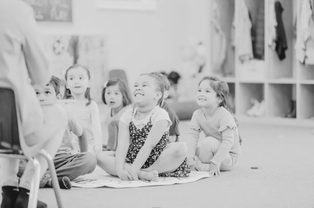 Group of young children sitting on a rug in a classroom, smiling and engaging with a person at the front.