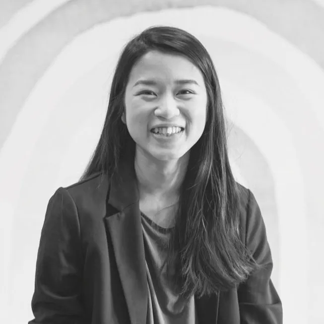 Black and white photo of a woman with long dark hair smiling, dressed in a blazer and blouse, against a circular background.