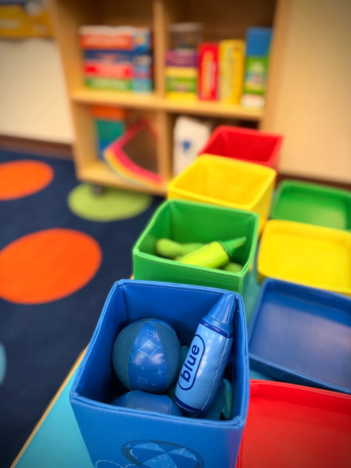 Colorful plastic bins filled with toys on a table in a playroom, blurred background with a bookshelf and colorful carpet with polka dots.