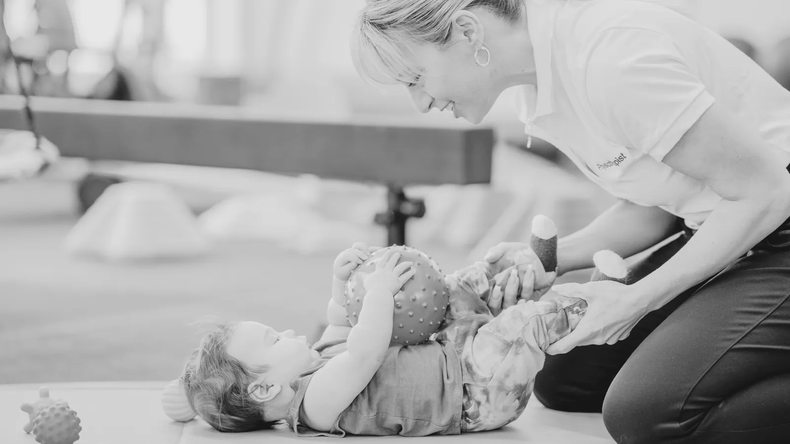 A child lying on the floor playing with a ball and other toys, while a woman, appearing to be a physical therapist, is helping the child by holding their legs and arms, engaging in a playful activity.