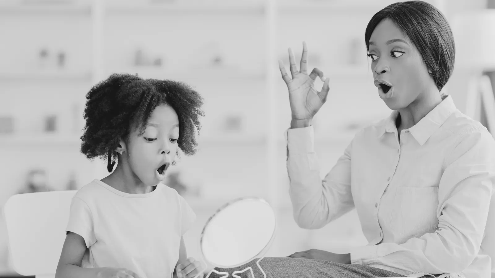 A woman and a young girl with curly hair are sitting at a table, looking surprised while the woman gestures with her hand near her face. The girl is holding a small mirror, and both are in a bright, modern room.