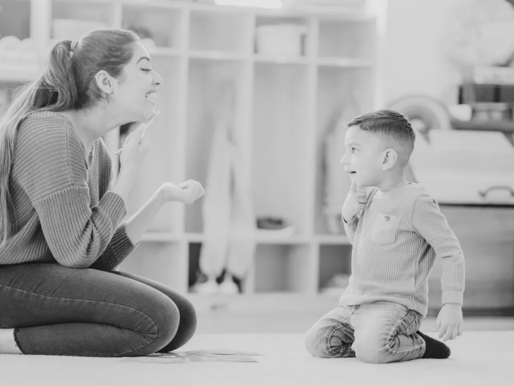 A young woman and a boy playing together indoors, with the woman kneeling on the floor and the boy sitting on his knees, both smiling and engaging with each other.