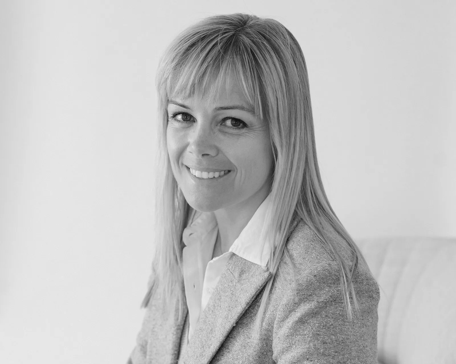 A black and white portrait of a woman with straight blonde hair, wearing a light-colored blazer and shirt, smiling and sitting in front of a plain background.