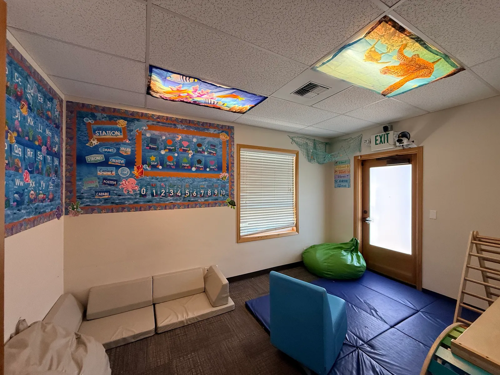 Part of a classroom with colorful educational decorations on the walls, including ocean-themed posters and a shelf with toys or educational materials. There are cushioned mats and bean bags on the floor, and a window with blinds.
