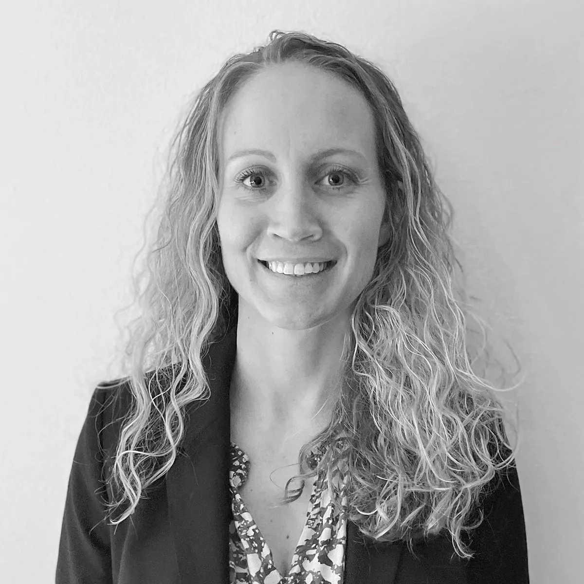 Black and white photo of a woman with curly hair, smiling, wearing a blazer over a patterned blouse, standing against a plain wall.