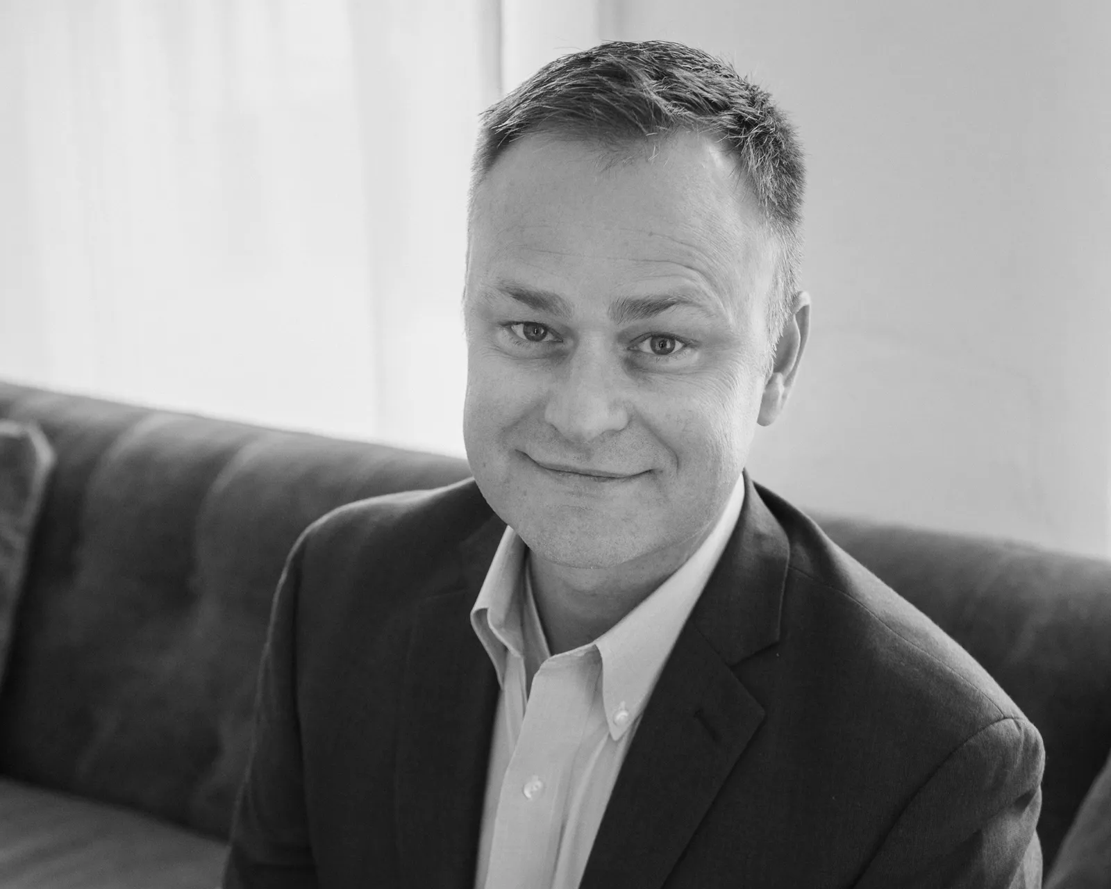 Black and white photo of a man with short hair, wearing a suit jacket and a collared shirt, sitting on a couch and smiling at the camera indoors.