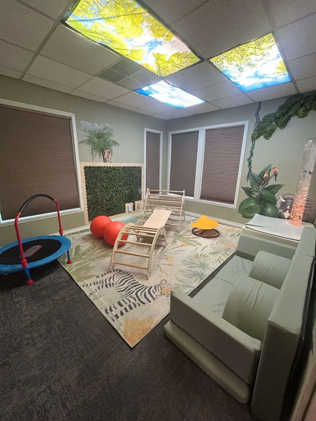 A room with a ceiling decorated with large ceiling tiles featuring outdoor sky and tree images, a gray wall with jungle and animal murals, windows with brown blinds, beige chairs, a small trampoline, exercise balls, a yellow container, and a white piece of furniture with a tall cylindrical lamp.