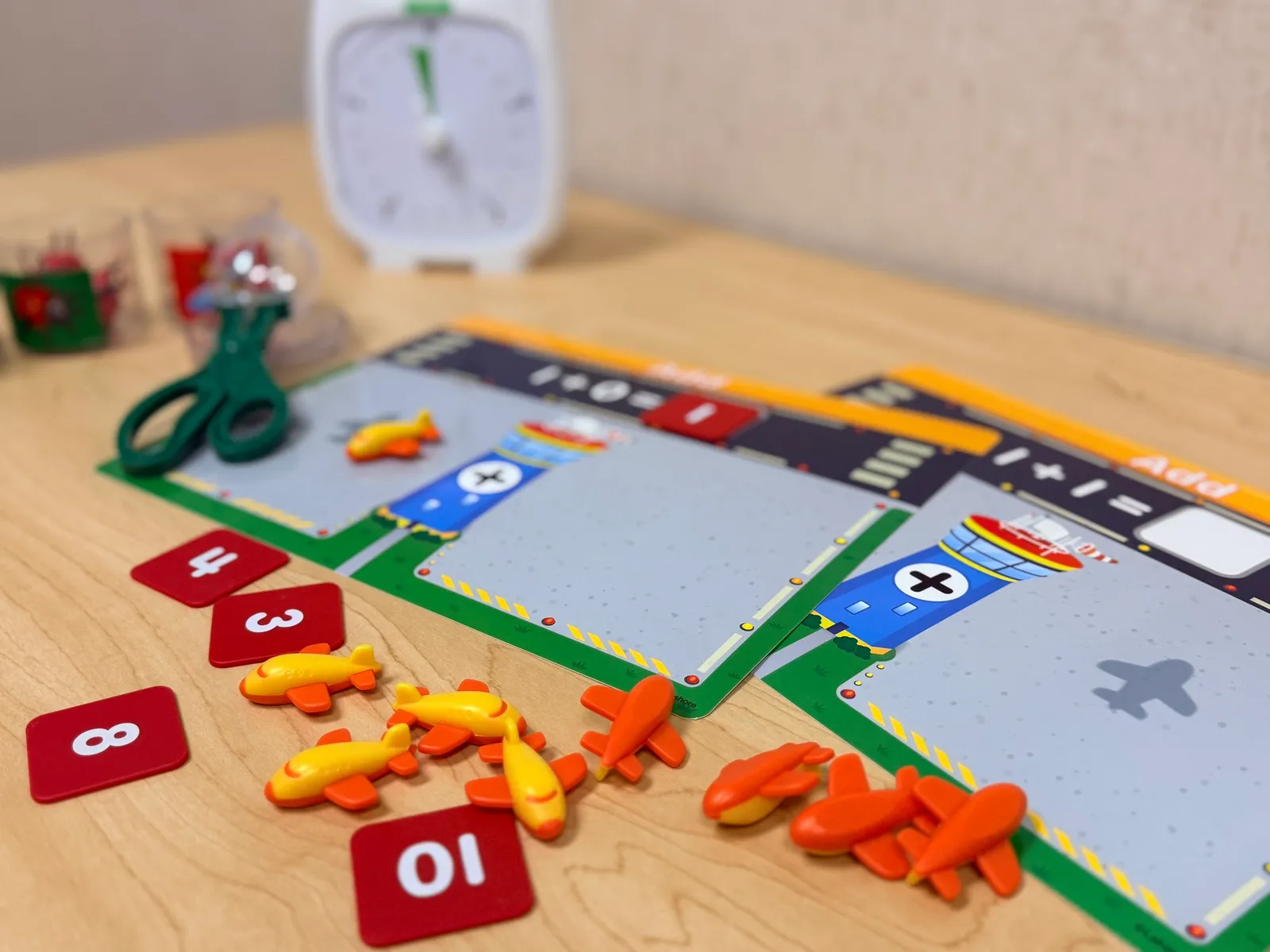 Close-up view of a Monopoly game in progress on a wooden table, with various game pieces including red property cards, colorful plastic tokens shaped like animals, and part of the game board visible, along with a blurred clock in the background.